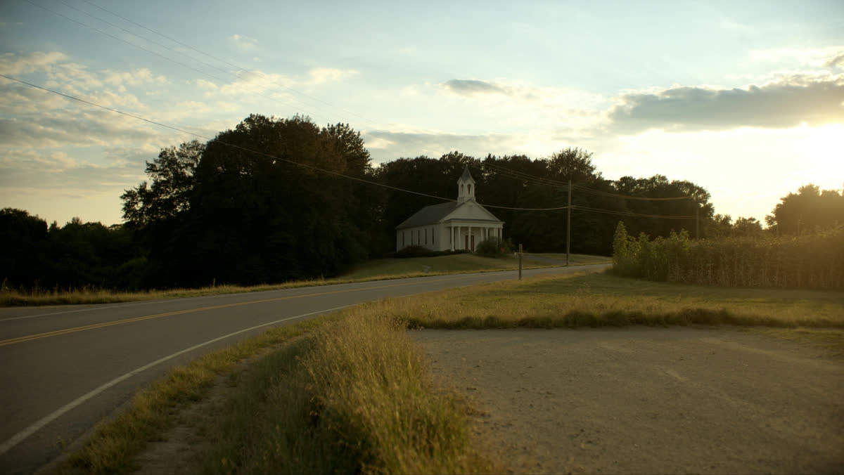 A quiet rural road curving toward a distant white church at golden hour