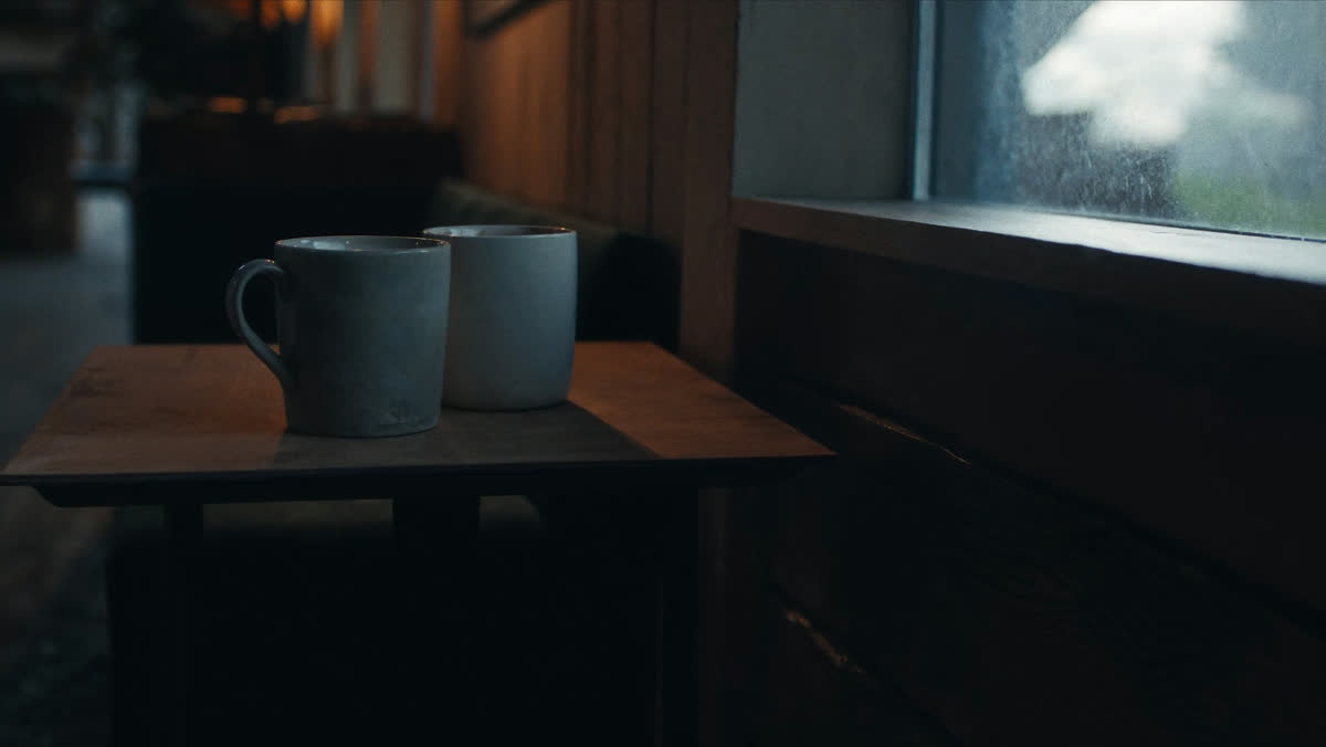 Two coffee mugs on a small table beside a rain-flecked window in a quiet corner