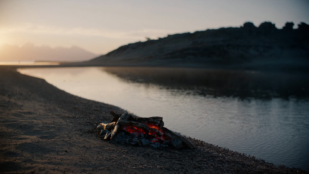 A small driftwood fire of glowing coals on an empty pebbled lakeshore at sunrise, a blackened iron pan with fish and a l